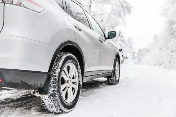 The Mufflerman, Mechanic Near Me A silver SUV with winter tire swaps is parked on a snowy road, surrounded by snow-covered trees. The focus is on the rear tire, highlighting its winter readiness, even during challenging times like COVID-19. The sky and surroundings are white and overcast.